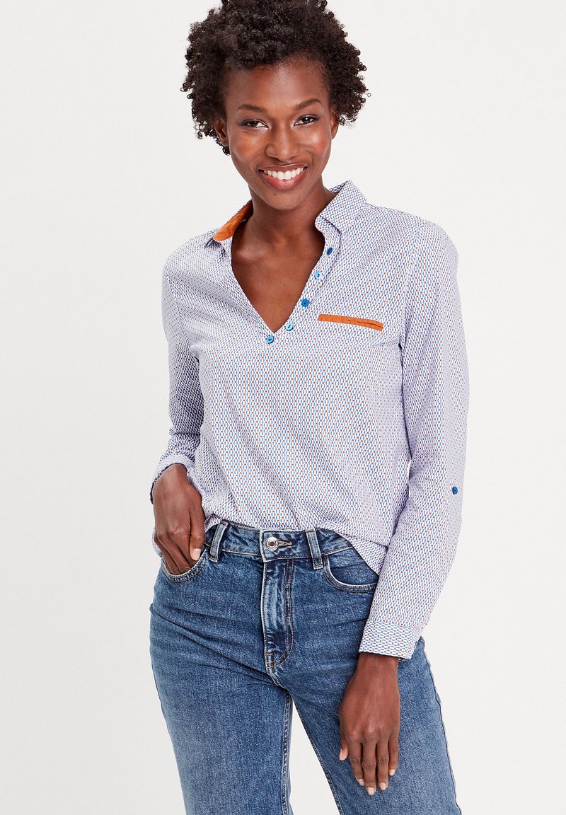 Chemise boutonnée blanche avec des motifs géométriques bleus, col orange contrastant et détails de poche. Assortie à un jean bleu taille haute.