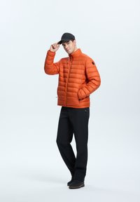Orange pufferjakke med lodrette sømme, lynlåslukning, sidelommer og en sort cap. Sammen med sorte bukser og robuste sko.