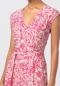 Blommig klänning i krämfärgad med livliga rosa blommönster, V-ringning, korta ärmar och knytning i midjan för en definierad silhuett.