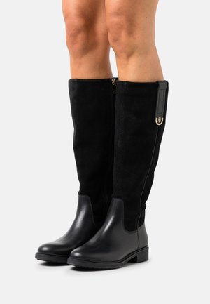 Stivali neri al ginocchio con punta in pelle e gambale in camoscio, indossati da una persona con gambe nude, con cerniera laterale e piccola targhetta con logo dorato.