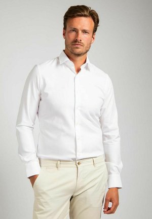 Man met lichtbruin haar, gekleed in een nauwsluitend wit overhemd en beige broek, staand met één hand in de zak tegen een neutrale achtergrond.