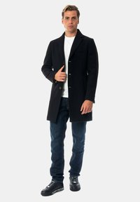 Cappotto di lana nera con colletto a punte, chiusura con bottoni e tasche laterali, indossato sopra una t-shirt bianca e jeans blu, abbinato a sneakers nere.