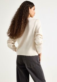 Maglione crema chiaro con vestibilità morbida, bordo a coste e maniche leggermente gonfie, abbinato a pantaloni a vita alta grigio scuro.