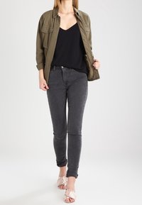 Žena v olivově zelené bundě přes černý top, spárovaná s tmavě šedými skinny džíny a růžovými sandály na otevřenou špičku s mašličkami.