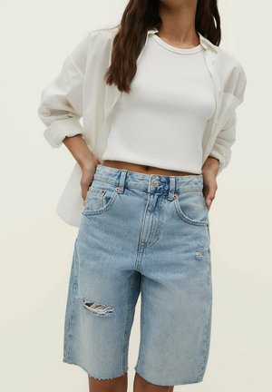 Kvinde iført lyseblå revne denimshorts, hvid ribbet crop top og en åben hvid skjorte, hænderne hvilende på hofterne.