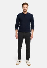Marinefarbener Strick-Polo-Pullover mit geripptem Textur, kombiniert mit dunkelgrauen, schmal zulaufenden Hosen und weißen Sneakern mit beigen Akzenten. Stehende Figur, die nach vorne blickt.