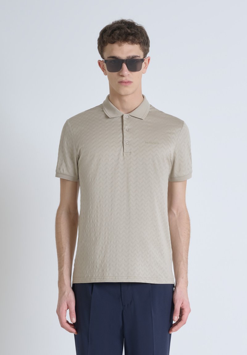 Camisa polo beige hecha de tejido texturizado con un sutil patrón de zigzag, que presenta un cuello clásico y mangas cortas. Gafas de sol negras puestas.
