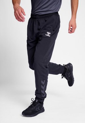 Schwarze Sporthose mit elastischem Bund, ausgestattet mit einem Hummel-Logo und Zickzackmuster an den Seiten. Dazu werden schwarze Schuhe getragen.