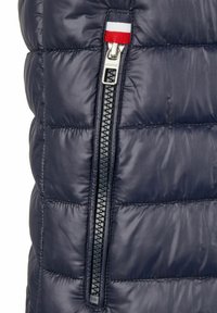 Marineblauer Steppweste mit einem gesteppten Design, silbernem Reißverschluss und einer rot-weiß-blauen Akzentlasche in der Nähe des Reißverschlusses.