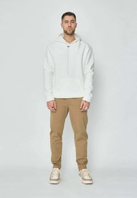 Un homme se tient face à la caméra, portant un hoodie blanc, un pantalon beige et des chaussures blanches, sur un fond clair et uni.