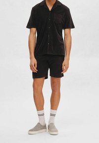 Camicetta nera a maniche corte con tasca, abbinata a pantaloni corti coordinati. Tessuto morbido, design del colletto classico, indossata con scarpe slip-on grigie e calzini bianchi a righe.