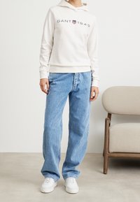 Sudadera crema con "GANT 1949" impreso en azul marino y burdeos, combinada con unos vaqueros de mezclilla sueltos y de color azul claro, y zapatillas blancas.