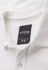 Biała kołnierzykowa koszula z czarną etykietą "ombre". Etykieta wskazuje rozmiar M (EUR 48). Posiada gładką fakturę i pojedyncze zapięcie na guzik.