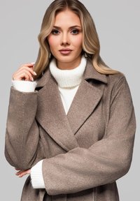 Vaaleanruskea villatakki, jossa on kauluskanta, vyötäröllä on vyö ja leveät hihat. Päällä valkoinen poolopaita.