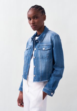 Γυναίκα με πλεξούδες, που φοράει μπλε τζιν μπουφάν με λεπτομέρειες με κουμπιά και λευκό παντελόνι, με φόντο απλό ανοιχτόχρωμο.