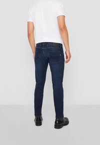 Man som står och visar baksidan av mörkblå slim-fit jeans, vit kortärmad skjorta och svarta läderskor på ljus bakgrund.