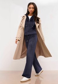 Sötétkék velúr tréningruha, zipzáros kapucnis felsővel és széles szárú nadrággal, amelyet strasszköves díszítések ékesítenek, bézs ballonkabát alatt viselve.