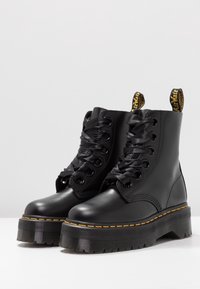 Botas de cuero negro hasta el tobillo con una suela de plataforma gruesa, punta redonda y cordones de satén negro. Presenta costuras amarillas y tiradores.