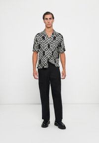 Man standing wearing black and white short-sleeve patterned shirt, black cargo pants, and black shoes against plain white background.