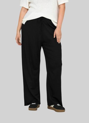Pantalons larges noirs avec une taille à cordon, fabriqués en tissu doux. La tenue est accompagnée de chaussures noires avec des lacets blancs et une semelle beige.