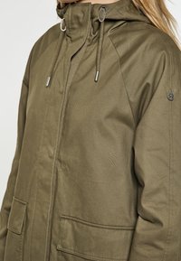 Giacca verde oliva di lunghezza media con zip e chiusura a scatto, dotata di cappuccio con cordino e due grandi tasche frontali. Tessuto liscio.