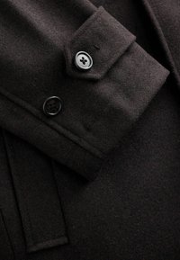 Rękaw ciemnobrązowego wełnianego płaszcza z dwoma czarnymi guzikami i kieszenią z klapką. Gładka tekstura z dopracowanymi detalami szycia.