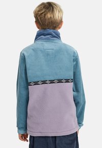 Giacca a pullover multi-colore con texture soffici in blu, viola e dettagli in corduroy, con una banda nera a motivi accentuata.