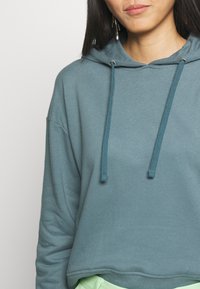 Sudadera corta con capucha en tejido verde azulado oscuro con un ajuste relajado, que presenta una capucha con cordón, dobladillo acanalado y un color de acento contrastante sutilmente visible.