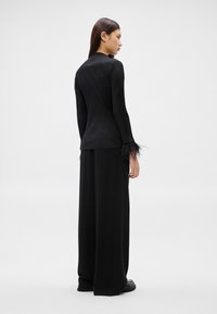 Žena stojaca v čiernom rebrovanom tričku s dlhým rukávom s pernatými manžetami a širokými čiernymi nohavicami na jednofarebnom bielom pozadí.