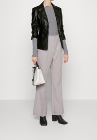 Žena v černém koženém saku, s dlouhým rukávem pruhovaným v černé a bílé barvě, lehkých šedých kalhotách, černých kotníkových botkách na podpatku, držící bílou kabelku.