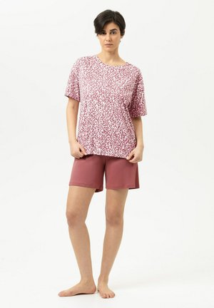 Vrouw staat op blote voeten en draagt een roze kortermouwshirt met patroon en effen roze korte broek tegen een effen witte achtergrond.