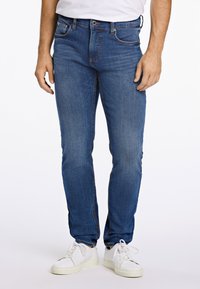 Uomo che indossa jeans blu aderenti, sneakers bianche e una maglietta bianca infilata nei pantaloni, in piedi davanti a uno sfondo semplice.