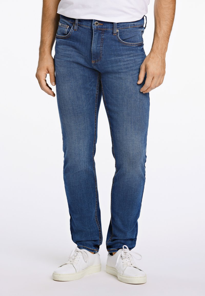 Uomo che indossa jeans blu aderenti, sneakers bianche e una maglietta bianca infilata nei pantaloni, in piedi davanti a uno sfondo semplice.