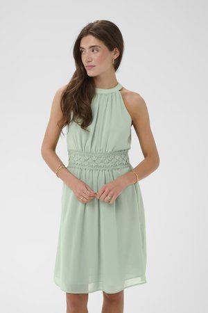 Frau trägt ein ärmelloses salbegrünes Kleid mit Spitzenbund, weitem Rock und Goldschmuck und schaut nach links.