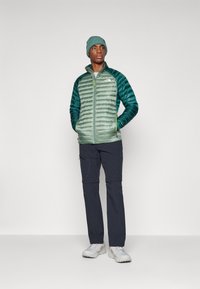 Groene geïsoleerde jas met horizontale naden, gecombineerd met donkere broeken en witte schoenen. Een bijpassende groene gebreide beanie wordt gedragen.