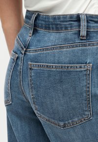 Gros plan d'une personne portant un jean en denim bleu avec des coutures visibles et une poche arrière, associé à un haut blanc.