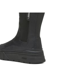 Musta nilkkuri, jossa on tyylikäs nahkapinta ja elastiset sivupaneelit. Varustettu paksulla, tekstuuripohjalla, jossa on brändätty yksityiskohta.