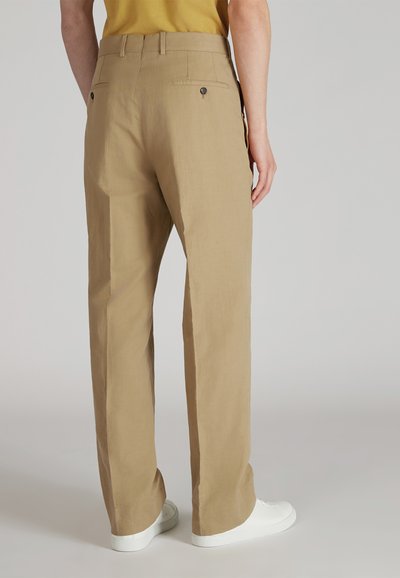 Personne portant un pantalon droit beige avec des poches arrière à boutons et des baskets blanches, se tenant devant un fond clair.