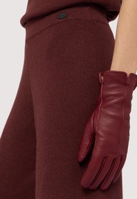 Bordowa skórzana rękawiczka o teksturowanym wykończeniu, umieszczona obok dopasowanej dzianiny z prążkowanym wzorem i metką z logo.