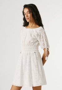 Γυναίκα με σκούρα μαλλιά που φοράει ένα λευκό φόρεμα με διχτυωτό σχέδιο, φουσκωτά μανίκια και εφαρμοστή μέση, στέκεται μπροστά σε ένα απλό ανοιχτόφωτο φόντο.