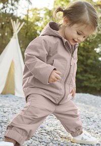Chaqueta rosa claro con cremallera y bolsillos, combinada con pantalones jogger a juego. Zapatillas blancas con cintas de velcro. Entorno al aire libre con grava.