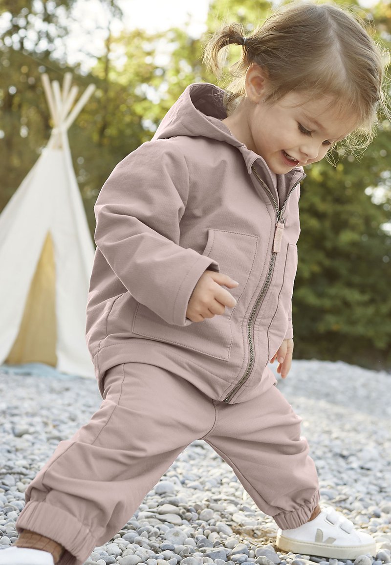 Chaqueta rosa claro con cremallera y bolsillos, combinada con pantalones jogger a juego. Zapatillas blancas con cintas de velcro. Entorno al aire libre con grava.