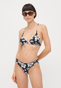 Melnā bikini komplekts ar baltu ziedu rakstu. Trīsstūra veida krūšturis ar plānām siksnām, atbilstošas zemas jostas bikses. Mīksts audums, minimālas segšanas dizains.