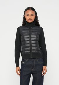 Μαύρο μπουφάν με επένδυση, φερμουάρ και ψηλό γιακά, με γυαλιστερό μπροστινό πάνελ και πλεκτά μανίκια. Συνδυάζεται με σκούρο τζιν.