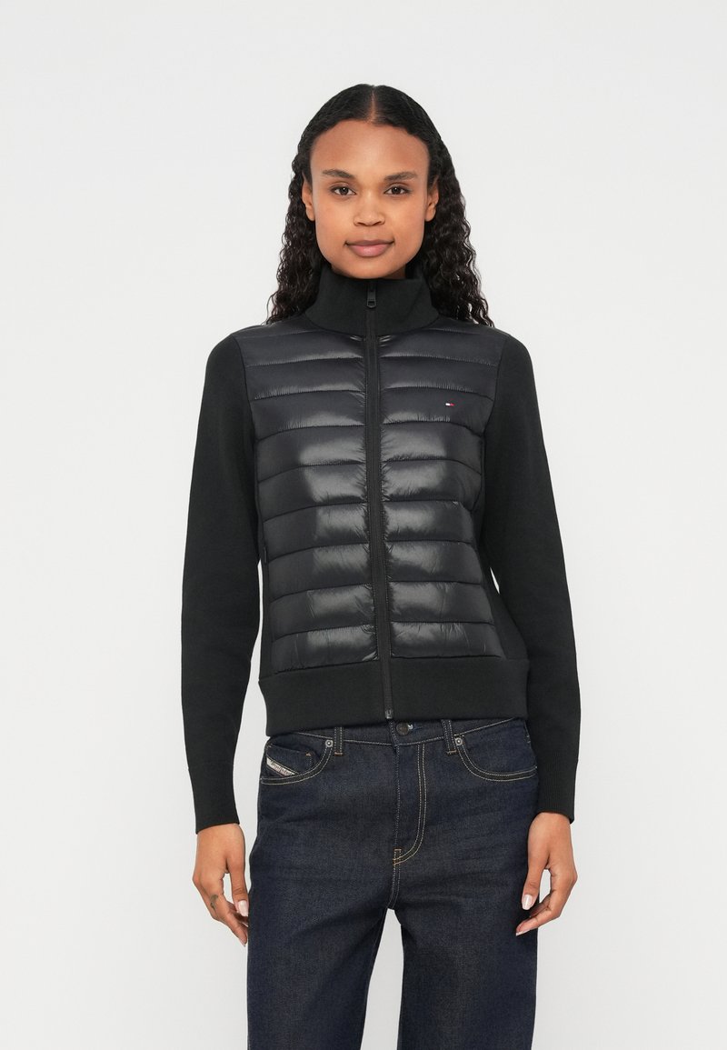 Μαύρο μπουφάν με επένδυση, φερμουάρ και ψηλό γιακά, με γυαλιστερό μπροστινό πάνελ και πλεκτά μανίκια. Συνδυάζεται με σκούρο τζιν.