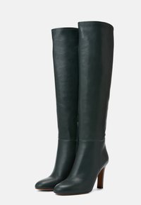 Bottes en cuir vert foncé montant jusqu'au genou, à bout rond et à talon en bois de hauteur moyenne, présentées côte à côte sur un fond clair.