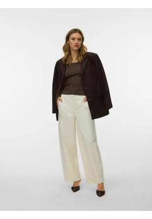 Žena stojaca s rukami v kapse, oblečená v tmavohnedom saku a zodpovedajúcom topu, krémových širokých nohaviciach a hnedých špicatých topánkach.