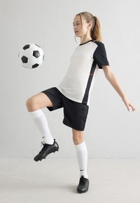 Jovem jogadora de futebol com uniforme preto e branco equilibrando uma bola preta e branca no joelho levantado contra um fundo branco simples.