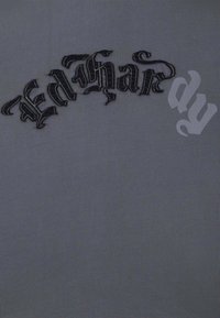 Harmaa T-paita, jossa on teksturoitu musta kirjonta "Ed Hardy" tyylitellyllä fontilla, ja vaatteessa on sileä ja joustava kangaspinta.