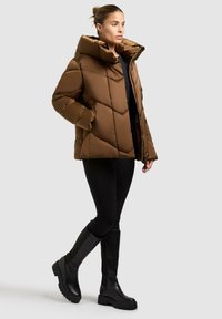 Giacca puffer marrone con design a rombi e cappuccio oversize, abbinata a leggins neri e stivali neri fino al ginocchio.